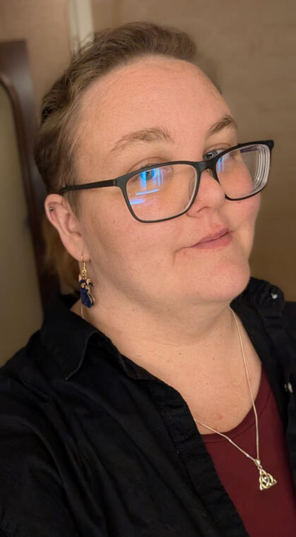 Me! A selfie of a white, blond-haired person with black framed glasses. They are looking at the camera with a small smile and are wearing a red shirt with a black over-shirt as well as a Celtic knot necklace and sailor moon earrings (specifically Luna).